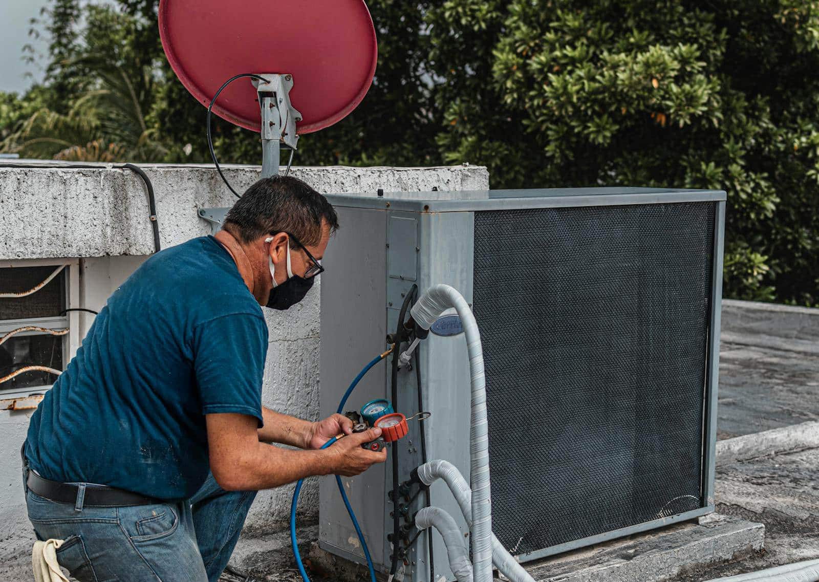 Technician repairing an air conditioner unit outdoors, wearing a facemask and using a manifold gauge.